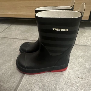 Svarta Tretorn stövlar med röd sula - Svarta stövlar från Tretorn med diskreta mörkgrå ränder och cool röd sula. Stövlarna har rund tå och är tillverkade i syntetmaterial som tål regn och slask. Perfekt för dig som vill ha stil även när det är blött ute.