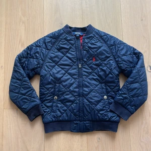 Mörkblå quiltad bomberjacka Polo Ralph Lauren - Quiltad mörkblå bomberjacka från Polo Ralph Lauren med röd broderad logga på bröstet. Jackan har dragkedja framtill, ribbade muddar och krage samt två fickor med tryckknappar. Insidan är fodrad i rött och materialet är polyester. Priset får gärna diskuteras.