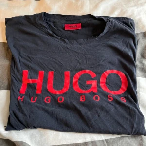 Marinblå t-shirt från Hugo Boss - Snygg Marinblå t-shirt från Hugo Boss med stor röd logotyp framtill. Klassisk rund hals och korta ärmar. Perfekt för dig som gillar stilrena märkesplagg med tydlig branding. Materialet är mjuk bomull som känns skönt mot huden.