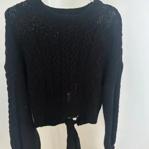 Svart stickad tröja med knyt från NA-KD - Svart stickad tröja från NA-KD med snyggt flätmönster och breda muddar. Tröjan har långa ärmar och en cool knyt-detalj baktill som man kan dra åt och knyta hur man vill.