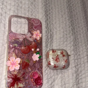 AirPods med fjärilsmönstrat skal + iPhone skal med 3D dekorationer - Säljer ett AirPods-fodral med vackert fjärilsmönster och ett iPhone-skal dekorerat med 3D-blommor, körsbär och glitter. Båda tillbehören är i fint skick, utan synligt slitage. Perfekt för dig som vill ha en personlig och färgglad look till din Apple-enhet.