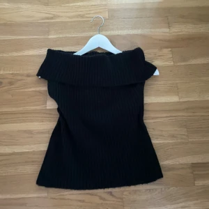 Svart ribbad offshoulder stickad topp - Snygg svart stickad topp med bred offshoulder-krage och ribbad struktur. Toppen är ärmlös från NA-KD