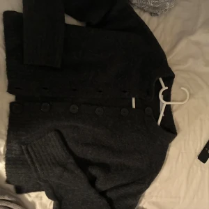 Mörkgrå stickad kofta med knappar - Mysig mörkgrå stickad kofta från lager157 med rund halsringning och stora svarta knappar framtill. Koftan har långa ärmar och breda ribbade muddar vid ärmslut och nederkant. Perfekt för lager-på-lager och chill höststil.