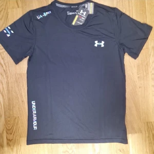 Svart träningst-shirt från Under Armour - Svart t-shirt från Under Armour med logga på bröstet och tryck på ärm och nederkant. Gjord i lätt och snabbtorkande polyester, perfekt för träning. Klassisk passform med korta ärmar och rund hals. Snygg och sportig stil för dig som gillar att röra på dig.