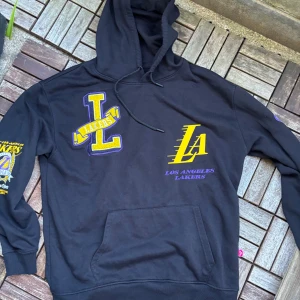 Svart Los Angeles Lakers hoodie - Svart hoodie med stor LA Lakers-logga i gult och lila på både fram- och baksidan. Tröjan har huva med snörning och en klassisk magficka. Feta tryck och broderade detaljer på bröstet och ärmen gör den riktigt sportig och unik.