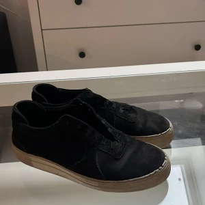Svarta sneakers från Arigato - Svarta sneakers från Arigato med klassisk låg profil och rund tå. Ovandelen är i mocka och skinn, med platt vit sula som har Arigato-logga undertill. Skorna har snörning men saknar skosnören på bilderna. Perfekt för dig som gillar stilrena och minimalistiska sneakers.
