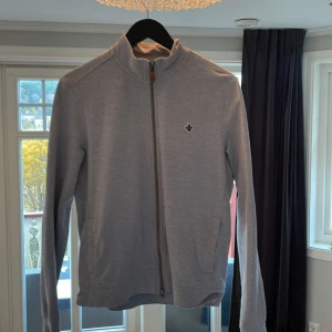 Grå ziptröja från Morris  - Snygg grå ziptröja från Morris i storlek S. Tröjan har hel dragkedja framtill, ståkrage och två fickor. Liten broderad logga på bröstet. Perfekt för dig som gillar stilrena och klassiska plagg.