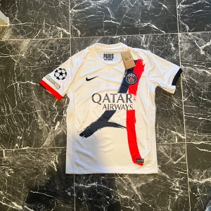 PSG Nuno Mendes Champions League tröja - Paris Saint-Germain fotbollströja med Nuno Mendes #25, Champions League-märke på ärmen och Qatar Airways sponsor. Vit bas med röd och mörkblå penseldrag, broderad PSG-logga och Nike swoosh. Tillverkad i lätt, ventilerande Dri-FIT-material.
