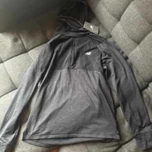 Svart löpar hoodie från New Balance - storlek M - Säljer en svart hoodie från New Balance i storlek M. Tröjan har huva med reflekterande band, tillverkad i ett lätt, snabbtorkande och andningsbart tyg. Perfekt för träning eller chill, med lång ärm och diskret logga på bröstet. Bröst ficka med dragkedja. Material: 95% återvunnen polyester, 5% elastan. Helt oanvänd med tagg kvar på tröjan. Skönt och värmande material på insidan. Pris kan diskuteras vid snabb affär. 