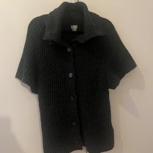 Svart stickad kofta med kort ärm - Säljer en svart stickad kofta med grov ribbad struktur och korta ärmar. Koftan har en bred krage och knäpps med fem svarta knappar framtill. Perfekt för dig som gillar chill och stilren look.
