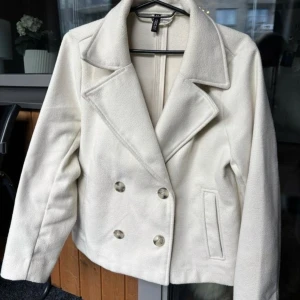 Vit kappa från H&M med knappar - Snygg vit kappa från H&M med dubbelknäppning och breda slag. Jackan har långa ärmar och två fickor framtill. Stilren och klassisk design som passar till många olika outfits. Materialet känns mjukt och har en ull-liknande struktur.