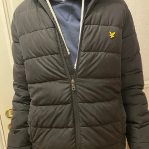 Svart pufferjacka från Lyle & Scott - Svart pufferjacka från Lyle & Scott med gul logga på bröstet. Jackan har huva, horisontella quiltade sektioner och dragkedja framtill. Perfekt för kalla dagar och har en clean, modern look som funkar till streetwear.