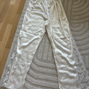 Vita sidenbyxor med spetsdetaljer - Säljer ett par vita, glansiga byxor i siden med elastisk midja. Byxorna har raka ben och är dekorerade med transparent spets längs sidorna, vilket ger en lyxig och romantisk vibe. Perfekta för dig som gillar mjuka material och snygga detaljer.