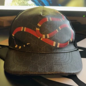 Gucci svart keps med ormprint - Snygg svart keps från Gucci med klassiskt GG-mönster och en röd orm som slingrar sig över framsidan. Justerbar rem med blå och röd detalj bak. Mesh på sidorna för extra ventilation. Perfekt för dig som vill sticka ut med lyxig streetstyle. Kepsen är storlek L och är i ett väldigt bra skick utan några skador. 