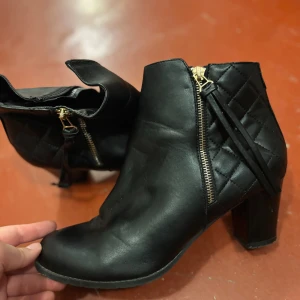 Svarta stövletter med dragkedja - Snygga svarta stövletter med quiltad detalj på sidan och guldfärgad dragkedja. Skorna har rund tå och stabil klack som ger extra höjd. Tillverkade i skinnimitation med stilren look, perfekta för att lyfta din outfit.