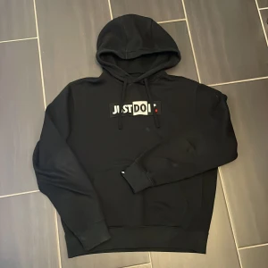 Svart Nike hoodie med Just Do It-tryck - Svart hoodie från Nike med broderat 'Just Do It.'-tryck på bröstet och liten Nike-logga på ärmslutet. Tröjan har huva med snörning, stor magficka och långa ärmar. Perfekt för dig som gillar streetwear och sportig stil.