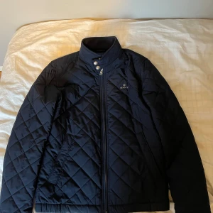 Mörkblå quiltad jacka från Gant - Snygg mörkblå quiltad jacka från Gant med hög krage, dragkedja och tryckknappar. Jackan har ett diskret Gant-logo på bröstet. Perfekt för dig som vill ha en stilren och klassisk look under höst och vår.