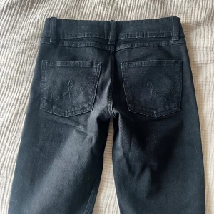 Svarta jeans från Gina Tricot - Svarta jeans från Gina Tricot med raka ben och klassisk femficksmodell. Jeansen har dubbla silverknappar framtill, dragkedja och snygga sömdetaljer på bakfickorna. Tillverkade i ett stretchigt bomullsmaterial för skön passform.