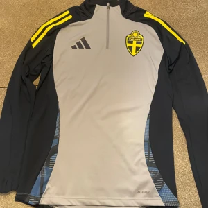 Svenska landslaget långärmad tröja Adidas - Slim fit långärmad fotbollströja från Adidas med Sveriges landslagsemblem på bröstet. Tröjan är grå med svarta och gula detaljer, har halv dragkedja och tillverkad i funktionsmaterial Aeroready. Snygga gula ränder på axlarna och svart Adidas-logga.