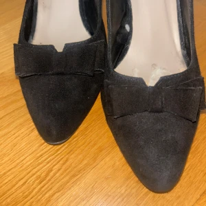 Svarta stilettklackar med rosett från Quazi - Snygga svarta stilettklackar från Quazi i mocka med spetsig tå och rosettdetalj framtill. Skorna har öppen häl med justerbar rem. De är köpta från Vinted och är lite slitna inuti som ni ser på bilderna, men annars i superbra skick! Klacken är ca 5-6cm 💘