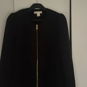 Svart kappa med dragkedja från H&M - Stilren svart kappa från H&M i storlek 36. Jackan har en rak passform, rundad halsringning och en snygg guldig dragkedja framtill. Dekorativa puffiga axlar. Långärmad och enkel design som passar perfekt till höst och vår. 