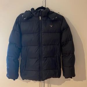 Svart pufferjacka från Gant 158/164 - Svart pufferjacka från Gant med huva och dragkedja framtill. Jackan är vadderad och har en klassisk quiltad look, perfekt för kalla dagar. Gant-logga på bröstet och praktiska fickor på sidorna. Passar dig som vill ha en stilren och varm vinterjacka.