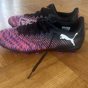 Svarta och lila Puma fotbollsskor - Säljer ett par grymma fotbollsskor från Puma med svart bas och coolt mönster i lila och rosa. Skorna har snörning, platt sula med dobbar och vit Puma-logga på sidan. Helt oanvända pga felköp storlek. 