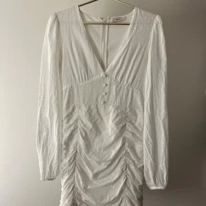 Vit kortklänning med v-ringning från Nelly - Supersnygg vit kortklänning från Nelly med v-ringad hals, långa ärmar och knappar framtill. Klänningen har ett diskret prickigt mönster och snygga rynkade detaljer längs sidorna. Dragkedja i ryggen för enkel påklädning. Perfekt för dig som gillar stilrena och trendiga plagg. Ser lite skrynklig ut nu men blir fin på. 