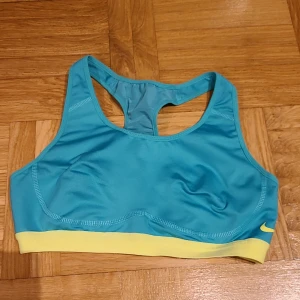Nike turkos sport-bh Dri-Fit S - Nike sport-bh i turkos med ljusgul resårkant. Modellen har brottarrygg och är tillverkad i Dri-Fit material som andas och transporterar bort fukt. Perfekt för träning och aktiva dagar. Snygg och sportig look med diskret Nike-logga framtill.