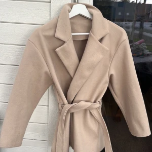 Beige kappa med bälte och krage - Stilren beige kappa med bred krage och knytskärp i midjan. Jackan har en klassisk omlottmodell och är tillverkad i ett mjukt ull-liknande material. Perfekt för dig som gillar en clean och trendig look. Jackan är köpt second hand men aldrig använd av mig. 