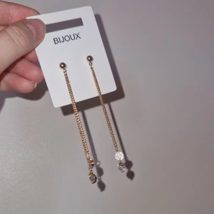 Långa guldiga örhängen med stenar från Bijoux - Snygga nya långa örhängen från Bijoux i guldig metall med kedjor och glittrande runda stenar längst ner. Perfekta för att ge din look lite extra bling! Örhängena har en stilren och elegant design som verkligen sticker ut.