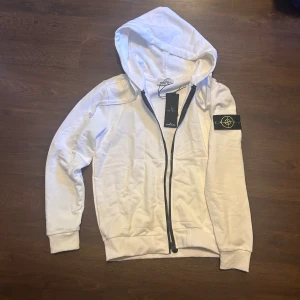 Vit hoodie från Stone Island storlek S - Helt ny Vit zip hoodie från Stone Island med klassisk svart och gul patch på vänster ärm. Tröjan har huva med snörning, hel dragkedja framtill och ribbade muddar. Tillverkad i mjuk bomull, perfekt för chill dagar. Snygg och enkel design med fickor framtill.