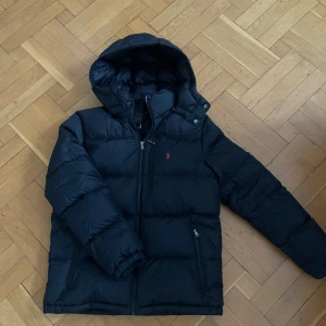 Svart pufferjacka från Polo Ralph Lauren - Svart pufferjacka från Polo Ralph Lauren med huva och dragkedja framtill. Jackan har två fickor med dragkedja och en liten röd logga på bröstet. Perfekt för kalla dagar och riktigt skön att bära.