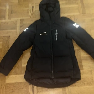 Svart dunjacka från Sail Racing XS - Svart dunjacka från Sail Racing med huva och reflekterande detaljer på ärmarna. Jackan är fylld med certifierat dun från Allied Feather + Down (500 fill power) och har dragkedja framtill. Vindtät, andas och är fluorkarbonfri. Perfekt för kalla vinterdagar.