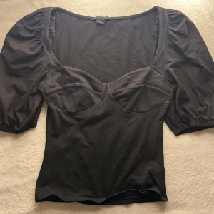 Svart topp med puffärmar från Monki - Svart topp från Monki med markerad byst, bred urringning och puffiga korta ärmar. Sitter figurnära och har en feminin siluett. Perfekt för dig som gillar trendiga detaljer och enkel stil.