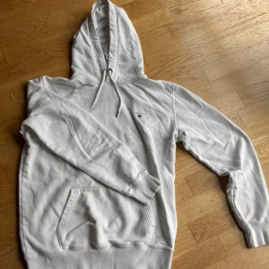 Vit hoodie från GANT med huva - Vit hoodie från GANT i storlek L (barnstorlek) med broderad logga på bröstet. Tröjan har huva med snörning, magficka och ribbade muddar vid ärmslut och nederkant. Mjuk insida i bomullsmix, perfekt för chill dagar. Klassisk och stilren design. Den är av väldigt bra kvalité och knappt använd. Nypris: 1250kr. Modellen är 173cm lång och väger cirka 70kg.