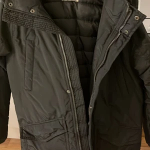 Svart parkas med fuskpäls - Svart parkas med huva och snygg fuskpäls runt kragen. Jackan har dragkedja, tryckknappar och flera praktiska fickor framtill. Perfekt för kalla dagar med sin vadderade insida och klassiska look.Vinterjacka från H&M till barn strl 146