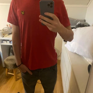 Röd t-shirt från Lyle & Scott - Röd t-shirt från Lyle & Scott med klassisk gul logga på bröstet. T-shirten har rund halsringning och är tillverkad i mjuk bomull. Personen på bilden är 178cm och väger 60kg. 
