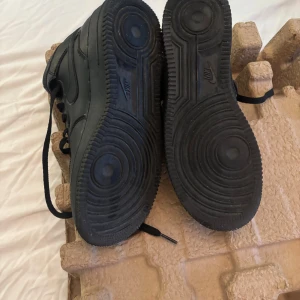 Nike Air Force 1 High svart skinn - Svarta Nike Air Force 1 High sneakers i skinn med klassisk siluett, snörning och kardborreband vid ankeln. Snygg helsvart look med perforerad tå och robust sula. Perfekta för dig som gillar streetstyle och vill ha en ikonisk sneaker i garderoben.