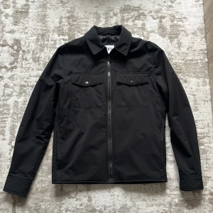 Svart vår/höstjacka med dragkedja - Stilren svart jacka från Zara med rak passform och klassisk krage. Jackan har två bröstfickor med lock och tryckknappar, samt hel dragkedja framtill. Materialet är syntet och ger en clean look som funkar till många outfits. Väldigt bra skick! 