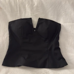 Svart korsett-topp med v-ringning - Snygg svart korsett-topp från NA-KD med markerad v-ringning framtill och smockade paneler på sidorna för extra komfort. Toppen är axelbandslös och har en croppad passform som ger en cool siluett. Perfekt att styla med höga jeans eller kjol. Den har en dragkedja på sidan så lätt att ta på sig!