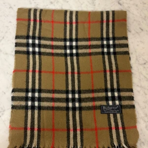 Rutig halsduk från Burberrys i ull - Klassisk rutig halsduk från Burberrys i 100% lammull. Färgerna är beige, svart, vit och röd med det ikoniska rutmönstret. Halsduken har fransar i ändarna och är tillverkad i England. Perfekt för att ge din outfit en snygg touch.