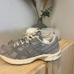 Grå Asics sneakers med mesh och mocka - Säljer ett par grå Asics sneakers med snygga meshpaneler och detaljer i mocka. Skorna har vita snören, rund tå och platt sula. Perfekt för dig som gillar sportig stil och neutrala färger. Ikonisk Asics-logga på sidan och bekväm vadderad insida. box ingår ej