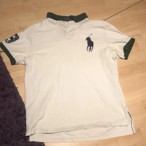 Vit piké från Polo Ralph Lauren - Vit pikétröja från Polo Ralph Lauren med gröna detaljer på krage och ärmslut. Stor broderad logga på bröstet och siffra 3 i rutigt tyg på ena ärmen. Klassisk passform och tillverkad i bomull. Perfekt för en sportig och stilren look.