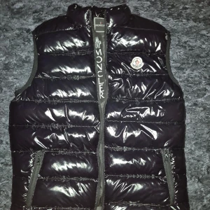 Svart dunväst från Moncler XL - Svart, glansig dunväst från Moncler i storlek XL. Västen har quiltad design, hög krage och dragkedja framtill med Moncler-logga på bröstet. Två praktiska fickor med dragkedja på sidorna. Perfekt för att addera en cool touch till din outfit. Skickar inom 2 dagar. Pris kan diskuteras