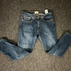 Blå jeans Grant regular fit 32/30 - Snygga blå jeans från Grant i regular fit med klassisk femficksdesign och kontrastsömmar. Modellen har raka ben och normal passform, med detaljer som bälteshällor och knappstängning. Perfekt för dig som gillar en enkel och tidlös jeansstil.