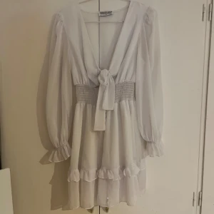 Vit kortklänning från Design By Amor - Supersöt vit kortklänning från Design By Amor med volangdetaljer och smock i midjan. Klänningen har långärmade transparenta ärmar, v-ringning med knyt framtill och volanger längst ner. Perfekt för dig som gillar romantisk och feminin stil.