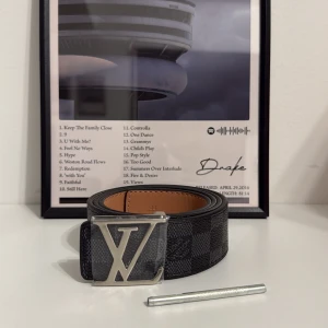 L och V rutig svart skinnbälte - Säljer ett L och V bälte i svart skinn med klassiskt rutigt Damier-mönster och en stor silverfärgad L och V-spänne. Insidan är ljusbrun och bältet har en lyxig vibe som verkligen sticker ut. Perfekt accessoar för att lyfta din outfit.