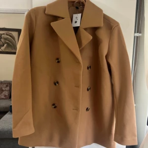 Klassisk beige skepparkavaj i ull - Snygg skepparkavaj i beige med dubbelknäppning och bred krage. Jackan har en rak passform och är tillverkad i ull, perfekt för kyliga dagar. Stilren och tidlös design som passar till det mesta.