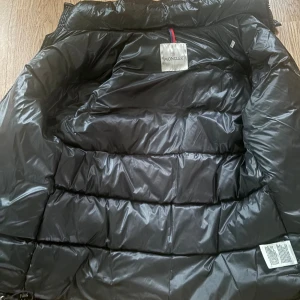 Svart Moncler dunjacka med huva - Snygg svart dunjacka från Moncler med glansig finish, huva och dragkedja framtill. Jackan har klassisk pufferstil med breda kanaler och Moncler-logga på ärmen. Perfekt för kalla dagar och riktigt stilren look.